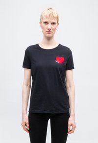 Άτομο με κοντά ξανθά μαλλιά που φοράει μαύρο μπλουζάκι με κόκκινη καρδιά και τη λέξη "AMOUR" στο στήθος, στέκεται μπροστά σε απλό φόντο.