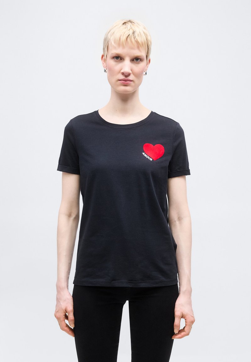 Άτομο με κοντά ξανθά μαλλιά που φοράει μαύρο μπλουζάκι με κόκκινη καρδιά και τη λέξη "AMOUR" στο στήθος, στέκεται μπροστά σε απλό φόντο.