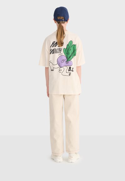 Personne portant un pantalon crème et un T-shirt ample crème avec un graphisme coloré de radis et du texte au dos, assortis à des chaussures blanches et une casquette marine.