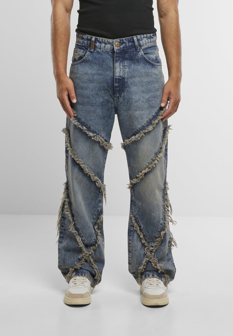 Mann trägt ausgewaschene blaue Jeans mit diagonalen ausgefransten Nähten und weiße Sneakers, steht vor einem schlichten hellen Hintergrund.