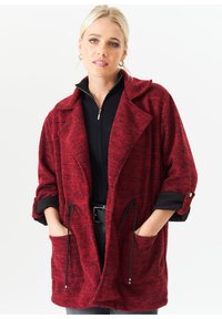 Red knit oversized coat with rolled sleeves, large pockets, and adjustable drawstring. Textured fabric with a subtle pattern.