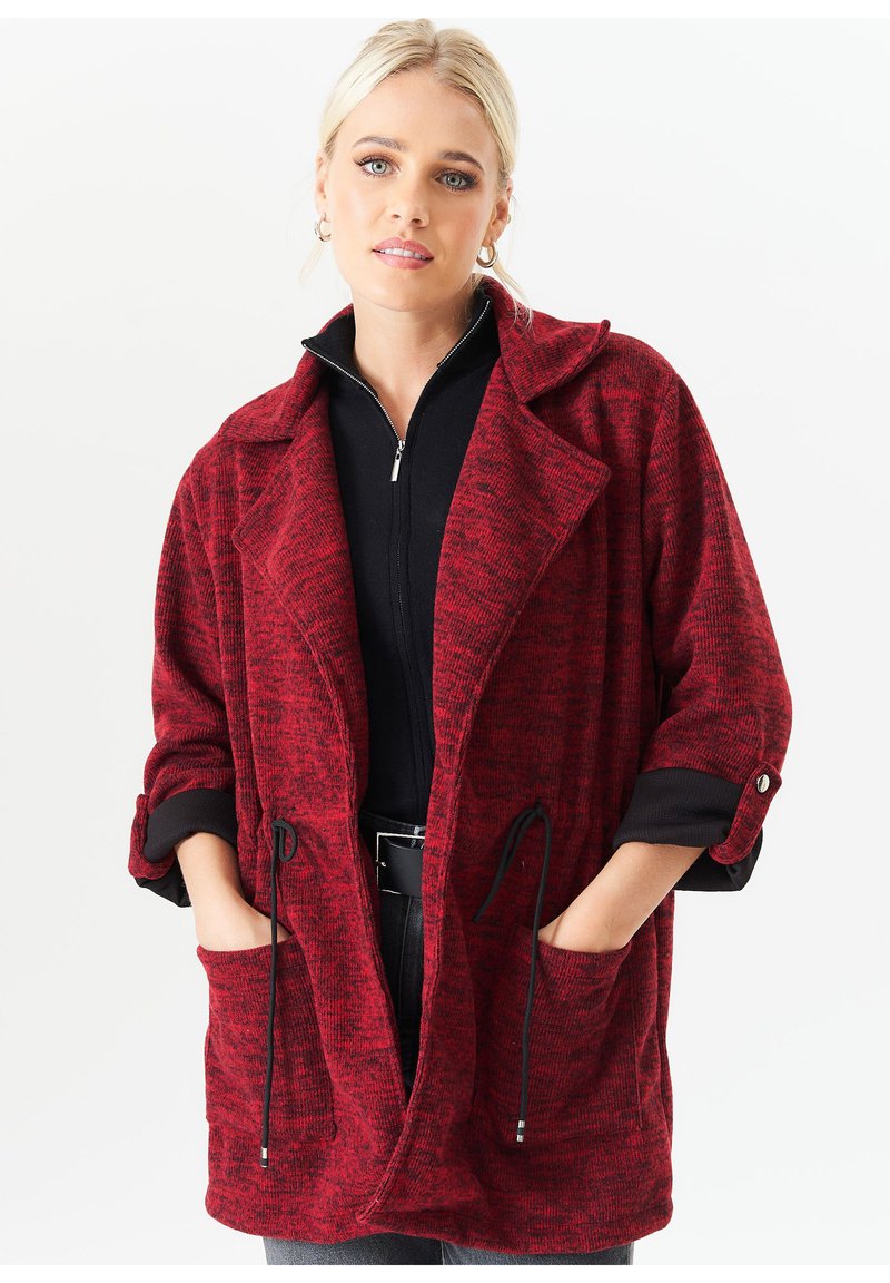 Red knit oversized coat with rolled sleeves, large pockets, and adjustable drawstring. Textured fabric with a subtle pattern.