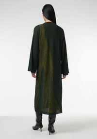 Lang groene cardigan met een ingewikkeld patroon van gele lijnen op de achterkant. Heeft wijde mouwen en valt onder de knieën. Soepel textuur.