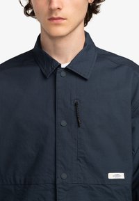 Navyblauwe knoop-jack met een textuurstof, een ritszak op de borst en een logolabel aan de onderzijde rechts.