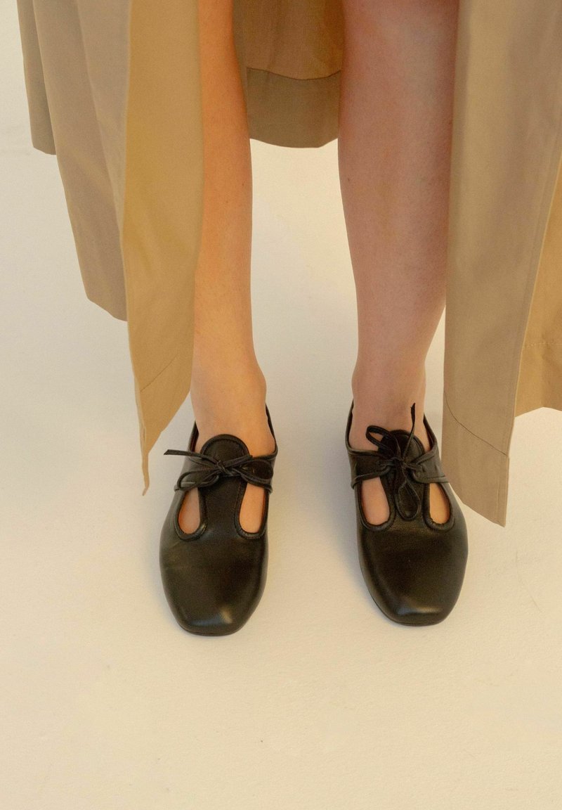 Jambes portant un pantalon beige et des chaussures en cuir noir à lacets avec des détails découpés sur un fond clair.