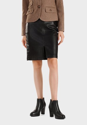 Donna che indossa un blazer marrone, una gonna in pelle nera al ginocchio con spacco frontale e stivaletti neri con tacco, in piedi su uno sfondo semplice.
