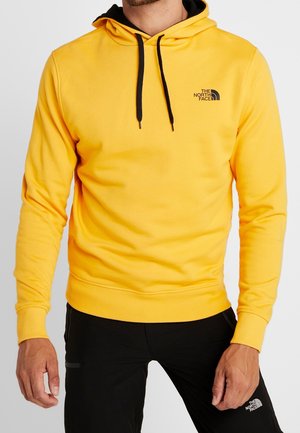 Homme portant un sweat à capuche jaune The North Face avec des cordons noirs et un pantalon noir avec le logo The North Face.