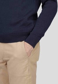 Pull marin avec des poignets et un ourlet côtelés, associé à un pantalon beige. Le pull a une texture lisse et une coupe classique.