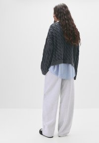 Kraftig marineblå kabelstriktrøje over en lyseblå stribet skjorte, parret med løse grå sweatpants og sorte slid-sko.