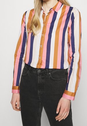 Donna che indossa una camicia a maniche lunghe con bottoni e righe verticali in arancione, rosa, blu navy e bianco, abbinata a jeans neri a vita alta.