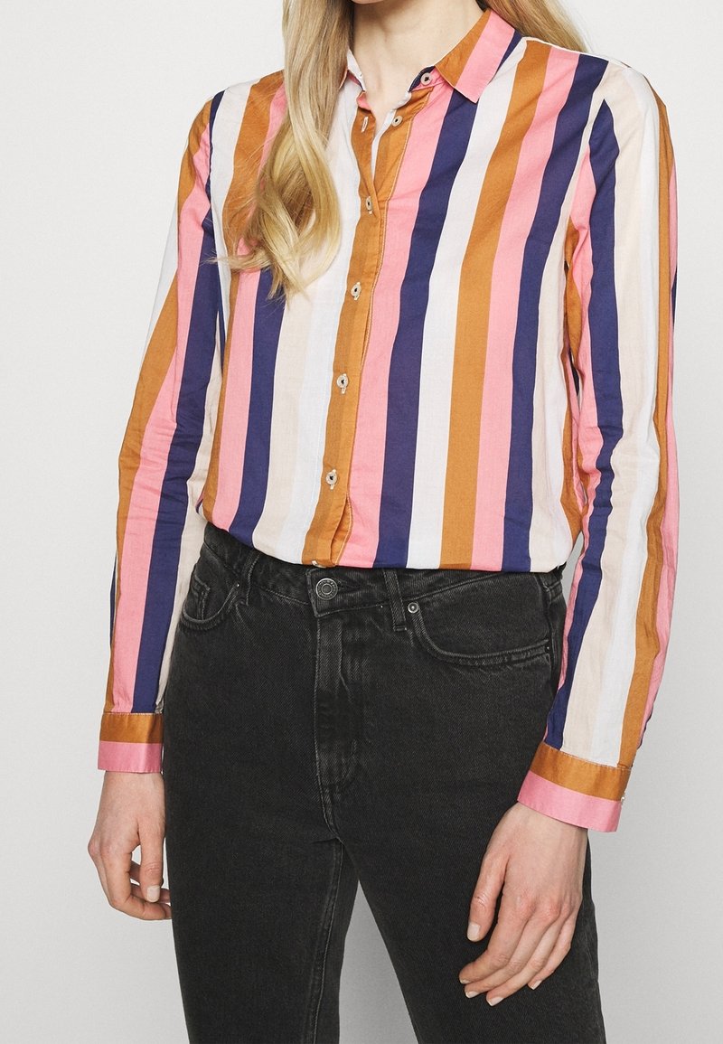 Femme portant une chemise à manches longues boutonnée avec des rayures verticales orange, rose, marine et blanche, associée à un jean noir taille haute.