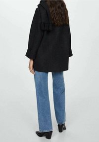 Kobieta w czarnym, oversize'owym płaszczu z szalem opadającym na jedno ramię, niebieskich prostych dżinsach oraz czarnych botkach na obcasie, stojąca tyłem.