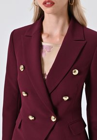 Blazer doppiopetto borgogna con bottoni dorati e rever affilati. Il tessuto appare liscio con una sartoria strutturata e tasche laterali.