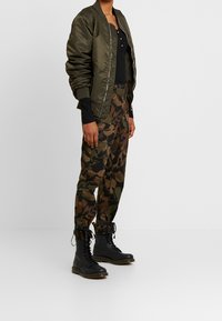 Oliivinvihreä bomber-takki, musta toppi, maastokuvioiset housut ja mustat nauhakenkäiset bootsit, joissa on paksu pohja, näkyvät saumat ja nauhat.