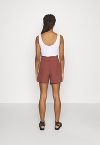 Hvid cropped tanktop, rustfarvede højlivede shorts og sorte sandaler, set fra ryggen mod en ensfarvet lys baggrund.