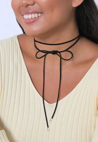 Crna choker ogrlica napravljena od mekane tkanine, sa mašnom sprijeda i zlatnim metalnim vrhovima na krajevima podesivih traka.