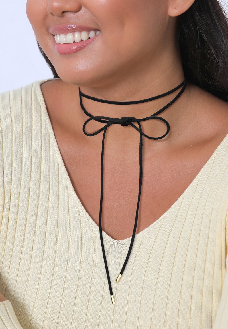 Crna choker ogrlica napravljena od mekane tkanine, sa mašnom sprijeda i zlatnim metalnim vrhovima na krajevima podesivih traka.