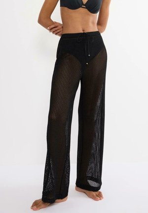 Frau trägt schwarze Netz-weitbeinige Hose mit Tunnelzug in der Taille über schwarzen Bikinihosen und einem schwarzen Bikini-Top, steht barfuß.