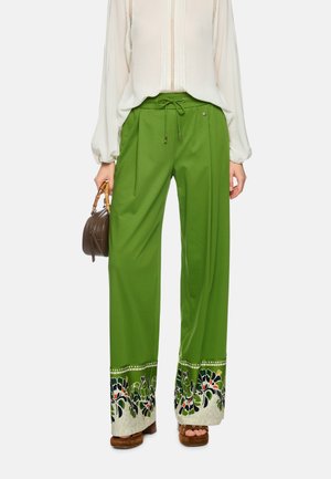 Frau trägt lockere grüne Hosen mit floralen Musterrändern, eine weiße langärmelige Bluse, braune Plateausandalen und hält eine braune Handtasche.