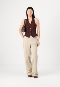 Gilet bordeaux senza maniche con bottoni, abbinato a pantaloni beige a righe larghe. Accessoriato con una piccola borsa a tracolla marrone.