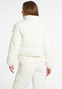 Jachetă albă pufoasă cu guler înalt, tiv canelat și manșete elastice. Textură netedă cu secțiuni quilted orizontale, fără hardware vizibil.