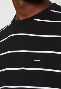Černé bavlněné tričko s horizontálními bílými pruhy, kulatým výstřihem a malou obdélníkovou cedulkou Calvin Klein na spodní části přední strany.