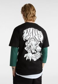 Osoba z blond włosami nosząca czarną koszulkę Vans, na plecach której znajduje się biały grafik przedstawiający groźnego tygrysa drapiącego kostki do gry.