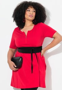 Vestido rojo de manga corta con cuello de cremallera, cinturón negro ceñido, bolsillos laterales y una textura suave; combinado con un bolso cruzado negro.