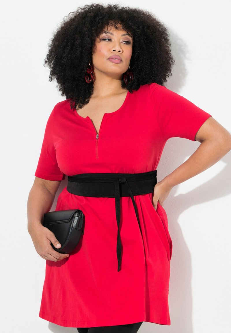 Vestido rojo de manga corta con cuello de cremallera, cinturón negro ceñido, bolsillos laterales y una textura suave; combinado con un bolso cruzado negro.