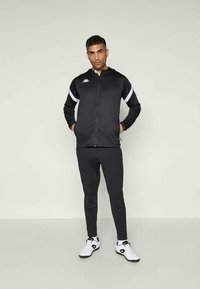 Svart zip-up jakke med hvite detaljer, sammen med svarte bukser og hvite joggesko. Jakken har en logo på brystet og en glatt tekstur.