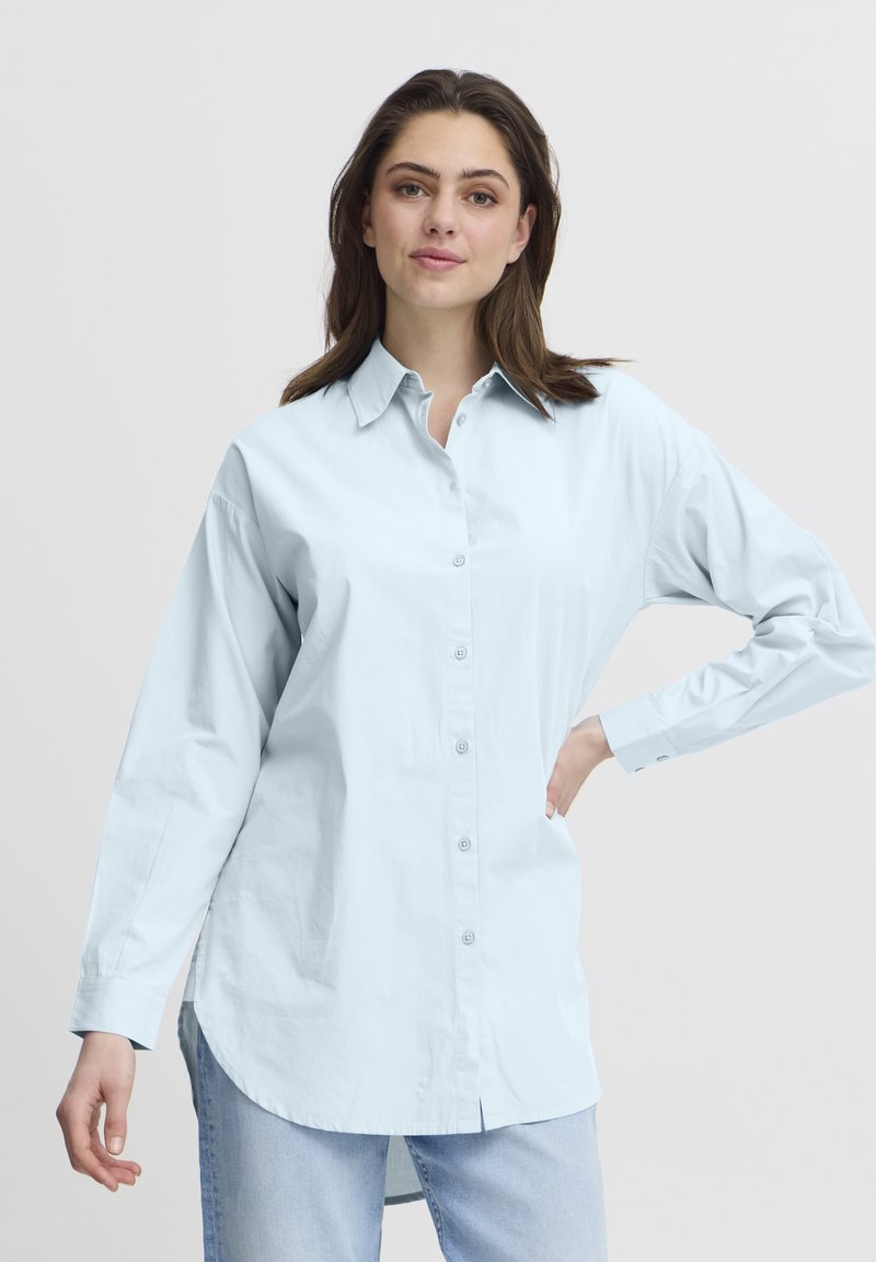 Camisa de botón de manga larga azul claro con collar, que presenta un corte recto y un dobladillo ligeramente curvado. Textura de tejido suave.