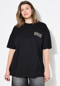 Camiseta oversized negra de algodón con mangas cortas, que presenta un texto impreso en multicolor en el área del pecho izquierdo.