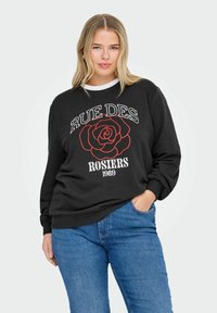 Mujer vistiendo un sudadera negra con una rosa roja y el texto "RUE DES ROSIERS 1989", combinada con unos vaqueros azules, con la mano en el bolsillo, fondo claro.