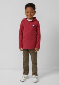 Ein lächelnder Junge, der einen roten Kapuzenpullover, braune Hosen und weiße Turnschuhe trägt, steht vor einem schlichten grauen Hintergrund.