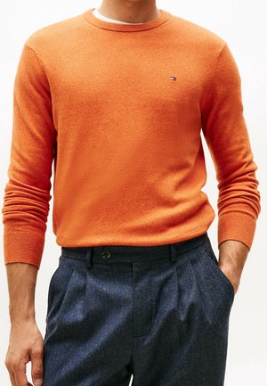 Mann trägt einen orangefarbenen Langarmpullover und dunkelblaue plissierte Hosen, mit einer Hand in der Tasche vor einfachem Hintergrund.