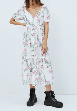 Frau trägt ein weißes Midikleid mit Blumenmuster und Puffärmeln sowie schwarze Schnürstiefel im Combat-Stil, steht mit einer Hand in der Hüfte.