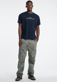 Marineblaues Baumwoll-T-Shirt mit "GUESS JEANS"-Logo; grüne Camouflage-Cargohosen mit Taschen; schwarze und braune Sneaker mit strukturiertem Obermaterial.
