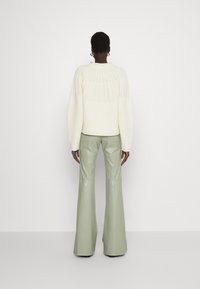 Neulepaita vaaleassa kerma-värissä, jossa on ribbikuvio, yhdistettynä vaaleanvihreisiin nahkaisiin levennettyihin housuihin. Suora taka-näkymä keskittyen vaatteiden yksityiskohtiin.