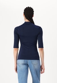 Navy blue, fitted top with short sleeves, high neck, smooth texture, and gathered fabric details at the back, paired with light blue jeans.