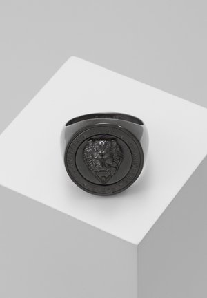 Anillo negro de metal con cabeza de león en relieve y la inscripción "GUESS LOS ANGELES" alrededor de la cara circular, exhibido sobre una superficie geométrica blanca.