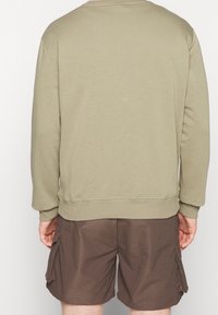Person klädd i en beige långärmad sweatshirt och bruna cargo-shorts, sedd bakifrån mot en vit bakgrund.