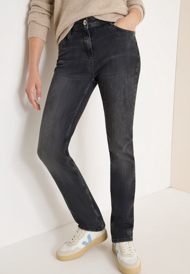 Personne portant un jean noir délavé, un pull beige et des baskets blanches avec des détails bleus, debout devant un fond clair uni.