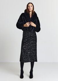 Schwarze Jacke aus Kunstpelz, kurz geschnitten, mit weitem Kragen, kombiniert mit einem schwarzen, mit Pailletten besetzten Midirock. Das Model trägt schwarze Strumpfhosen und hochhackige Schuhe.