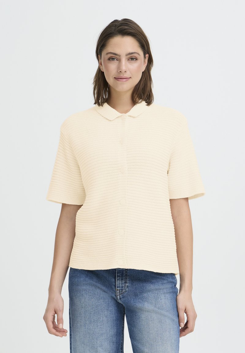 Femme aux cheveux bruns jusqu'aux épaules portant une chemise à manches courtes texturée crème et un jean bleu, se tenant devant un fond blanc.