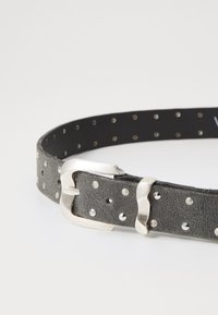 Fekete bőrszíj ezüstfém csattal, szabályosan elhelyezett kerek lyukakkal a hevederen a beállításhoz. Texturált felület részletek.