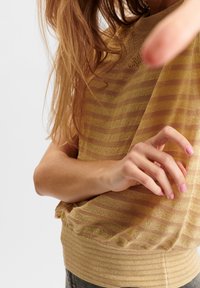 Lekki dzianinowy top w żółte i brązowe poziome paski, z prążkowanym dołem i fakturowanym wykończeniem. Ręka umieszczona z przodu, z różowymi paznokciami.