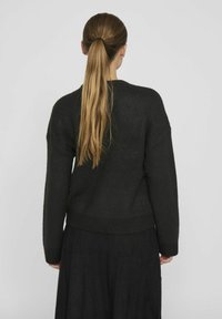 Tmavě zelený, oversized svetr vyrobený z měkké, strukturované tkaniny s kulatým výstřihem a ribbaným lemem. Nosený s dlouhou černou sukní.