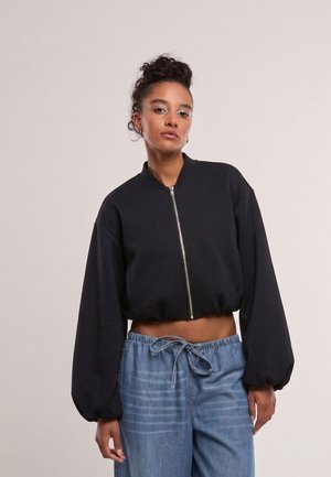 Femme aux cheveux bouclés en chignon haut portant une veste noire courte zippée et un jean bleu ample à cordon, sur un fond uni.