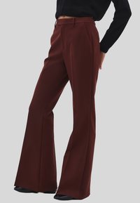 Persona che indossa pantaloni a vita alta e gamba larga color bordeaux con tasche laterali e scarpe nere, su uno sfondo grigio.