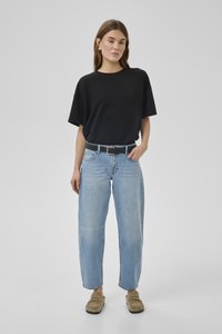 Sieviete, kas valkā melnu pārāk lielu T-kreklu, gaiši zilas augsta jostasvietas džinsas ar melnu jostu, bēšas loafer kurpes un pērļu auskarus, stāvot ar vienu roku kabatā.